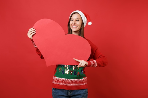 Sonriente joven Santa con sombrero de Navidad apuntando con el dedo índice en el corazón rojo en blanco vacío en manos aisladas sobre fondo rojo. Feliz año nuevo 2019 celebración concepto de fiesta navideña. Simulacros de espacio de copia.