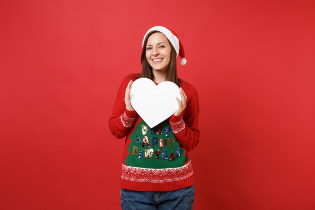 Sonriente a joven Santa en suéter de punto, sombrero de Navidad con corazón vacío en blanco blanco aislado sobre fondo rojo brillante. Feliz año nuevo 2019 celebración concepto de fiesta navideña. Simulacros de espacio de copia.