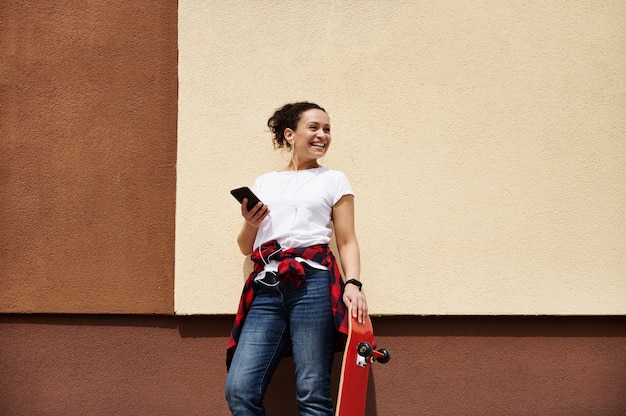 Sonriente joven skater con patineta roja de madera sosteniendo smartphone disfrutando de escuchar música mientras posa al aire libre cerca de la pared