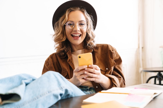 Foto sonriente joven con sombrero utilizando teléfono móvil con piernas sobre la mesa mientras estudia en casa