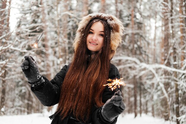 Sonriente joven sosteniendo bengala en la mano de pie en el bosque de invierno mirando a la cámara al aire libre