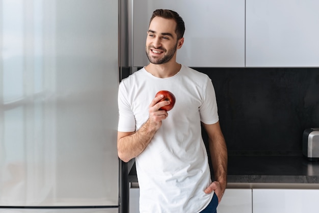 Sonriente joven sosteniendo una manzana mientras está de pie en la cocina de casa