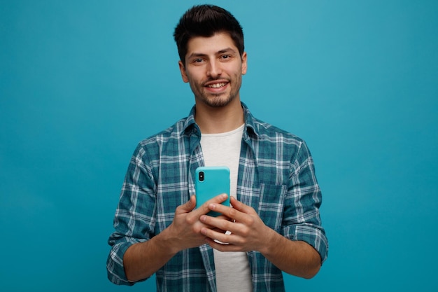 Sonriente joven sosteniendo teléfono móvil mirando a la cámara aislada sobre fondo azul.