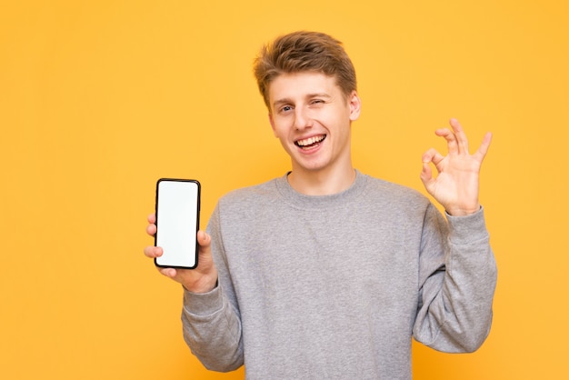 Sonriente joven sostiene un teléfono inteligente con una pantalla en blanco y muestra un signo de OK en un amarillo