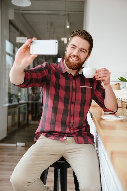 Foto sonriente joven tomando selfie y sosteniendo la taza de café