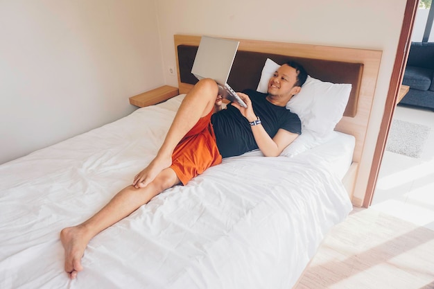 Sonriente joven trabajando desde la cama Acostado mientras revisa la computadora portátil