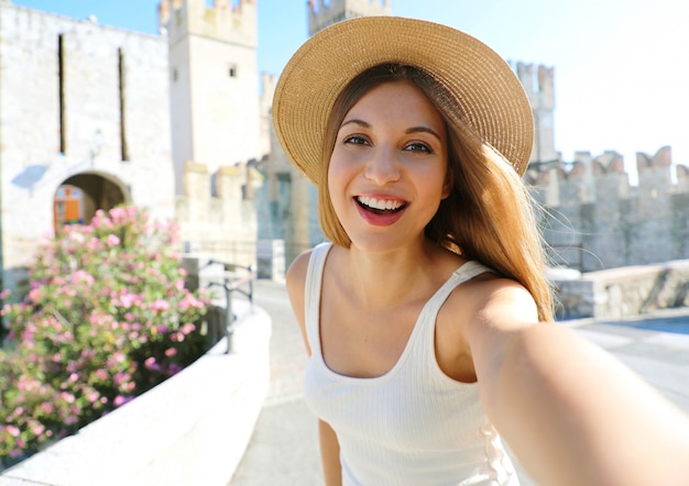 Sonriente joven turista toma autorretrato en Sirmione, Italia. Hermosa chica de moda toma una foto selfie con el castillo de Sirmione en el fondo, el lago de Garda, Italia.