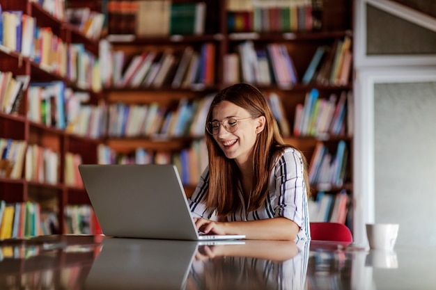 Sonriente joven universitaria atractiva sentada en la biblioteca y usando la computadora portátil. Ella está escribiendo la tarea.