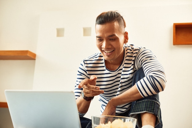 Sonriente joven vietnamita comiendo papas fritas y viendo una película de comedia en la pantalla del portátil