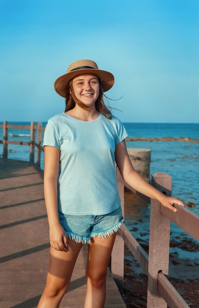 Sonriente jovencita de pie sobre la pasarela de madera junto al mar al atardecer, vestida con camiseta azul claro y sombrero de paja. Concepto de viaje de verano
