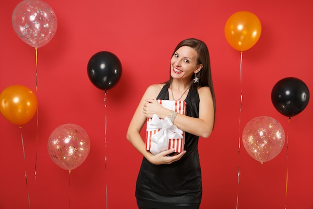 Sonriente a jovencita vestida de negro celebrando, abrazando con caja roja con regalo, presente en globos de aire de fondo rojo brillante. Día de la mujer, feliz año nuevo, concepto de fiesta de vacaciones de maqueta de cumpleaños.