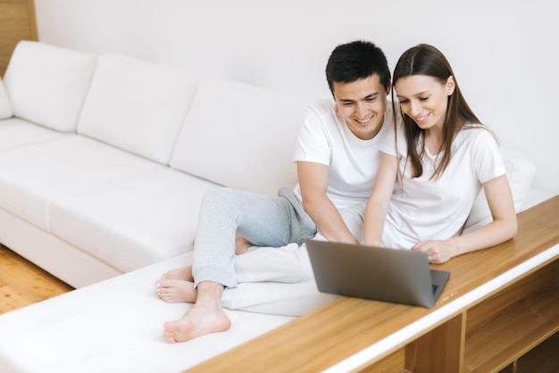 Sonriente y linda pareja joven está sentado en un sofá blanco y usa una computadora portátil en la luminosa sala de estar, compras en línea. Feliz pareja usa portátil sentado en un cómodo sofá en casa