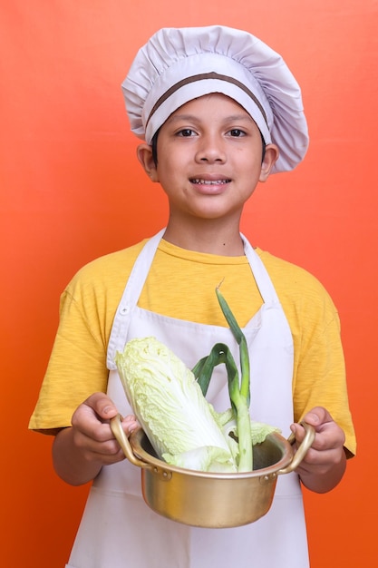 Foto sonriente y lindo niño asiático con uniforme de chef sosteniendo una sartén de verduras frescas