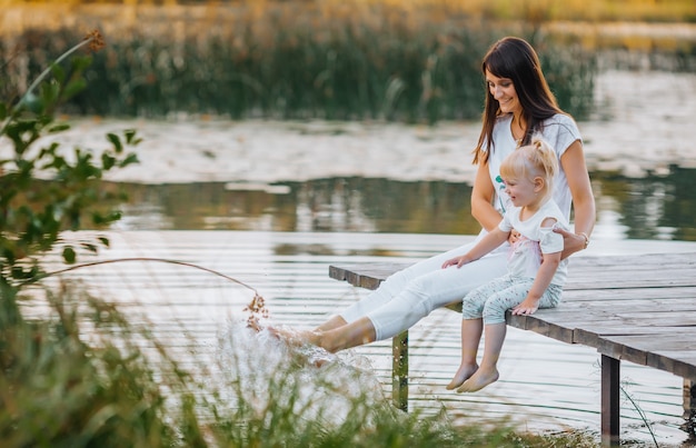 Sonriente madre e hija sentada en el muelle cálido día de otoño colgando sus piernas en el agua al atardecer