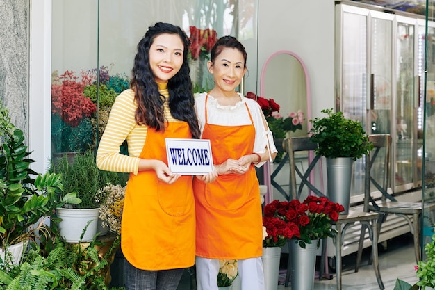 Sonriente madre e hija vietnamita en delantales naranja abriendo floristería