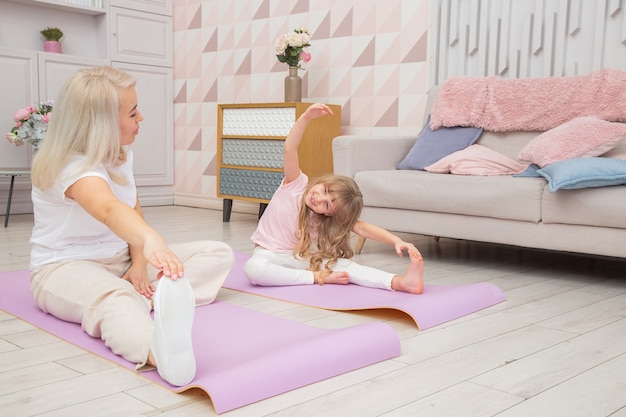 Sonriente madre rubia en estera de yoga con linda hijita preescolar juguetona hacer varios ejercicios