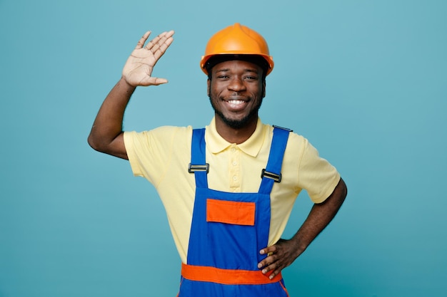 Sonriente mano levantada joven constructor afroamericano en uniforme aislado sobre fondo azul.