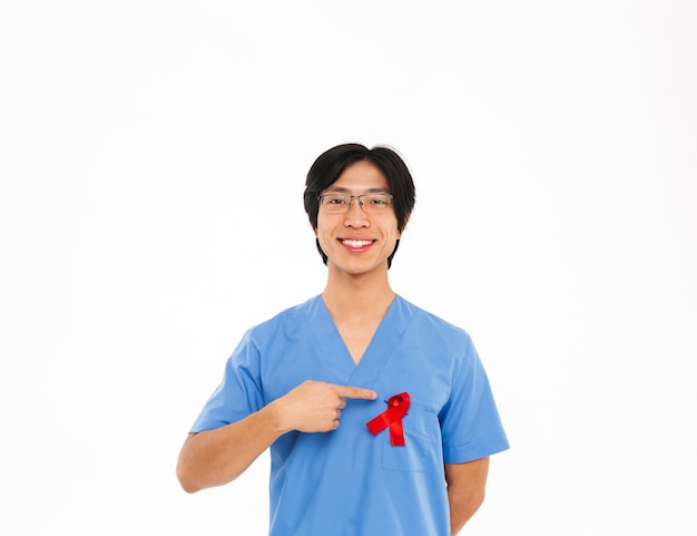 Sonriente médico asiático hombre vestido con uniforme con cinta roja que se encuentran aisladas sobre la pared blanca