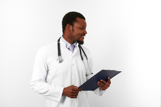 Sonriente médico barbudo negro con túnica blanca y estetoscopio tiene historial médico en el portapapeles
