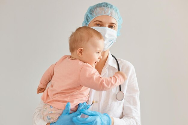 Sonriente médico pediatra satisfecho con gorro médico, mascarilla quirúrgica, bata y guantes de goma, de pie con una niña pequeña en sus brazos, mirando a la cámara, expresando emociones positivas.