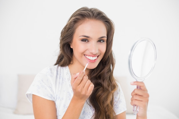 Sonriente morena atractiva aplicando brillo de labios y sosteniendo espejo