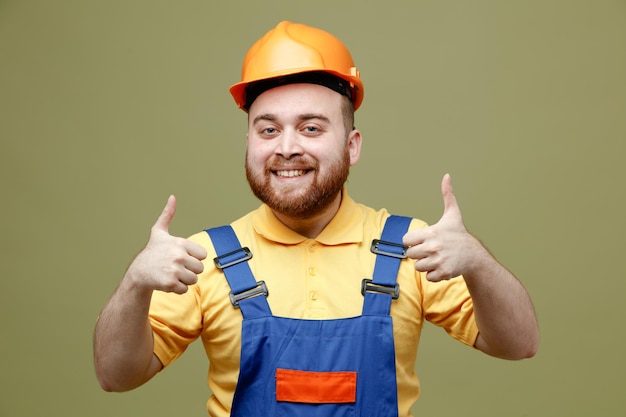 Sonriente mostrando pulgares joven constructor hombre en uniforme aislado sobre fondo verde