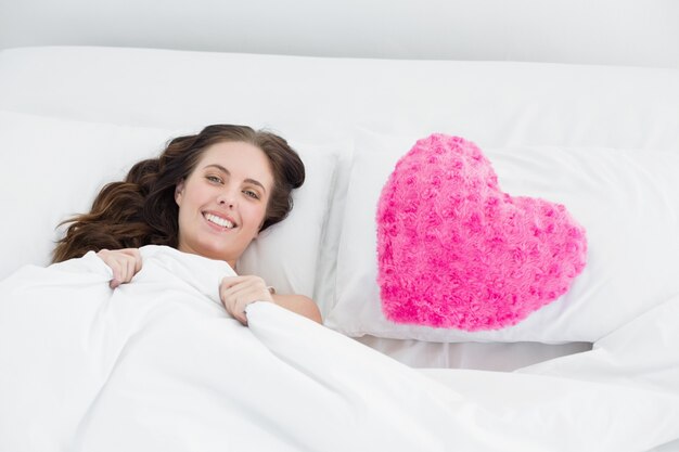 Sonriente mujer acostada en la cama con la almohada en forma de corazón