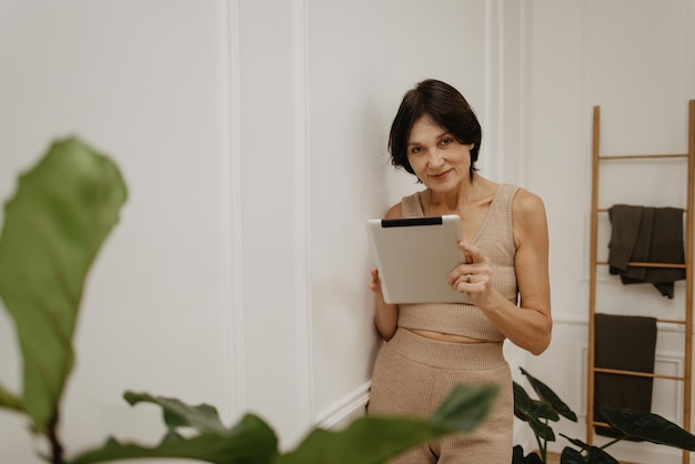 Sonriente mujer adulta caucásica mirando a la cámara sosteniendo la tableta mientras está de pie contra la pared blanca Estilo de vida de ocio y concepto de belleza