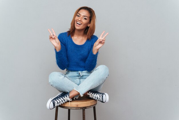 Foto sonriente mujer africana en suéter y jeans sentado en una silla y mostrando la paz. fondo gris aislado