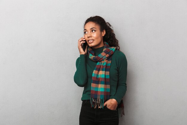 Sonriente mujer africana vistiendo abrigo de pie, mediante teléfono móvil