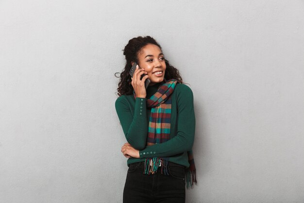 Sonriente mujer africana vistiendo abrigo de pie, mediante teléfono móvil