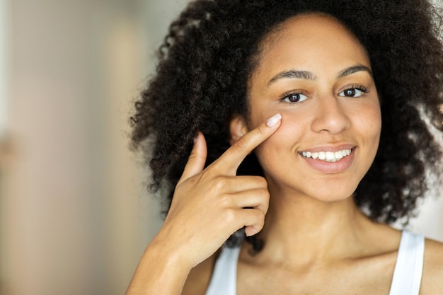 Sonriente mujer afroamericana aplicando crema hidratante facial mientras mira la cámara.