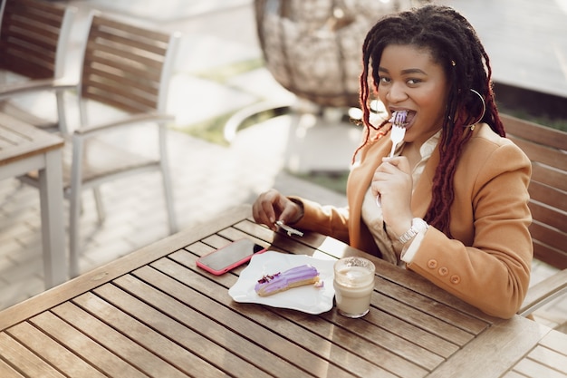 Sonriente mujer afroamericana bebiendo café y comiendo postre en la cafetería al aire libre