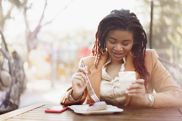 Sonriente mujer afroamericana bebiendo café y comiendo postre en la cafetería al aire libre