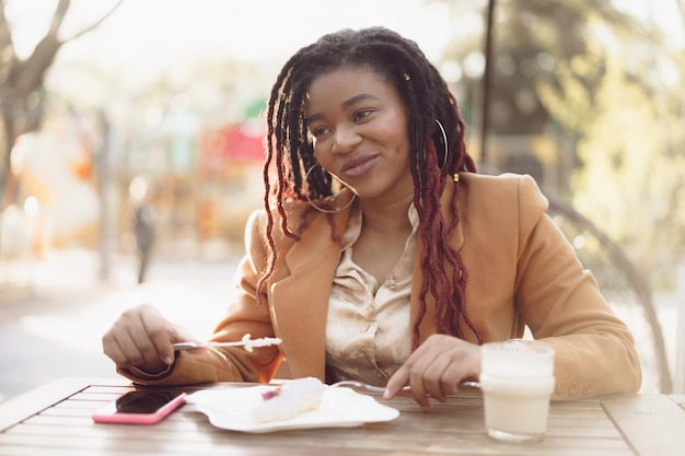 Sonriente mujer afroamericana bebiendo café y comiendo postre en la cafetería al aire libre