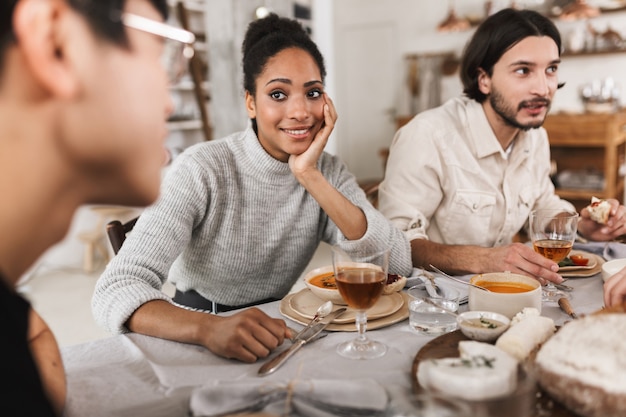 Sonriente mujer afroamericana con cabello rizado oscuro sentado en la mesa apoyándose en la mano pensativamente