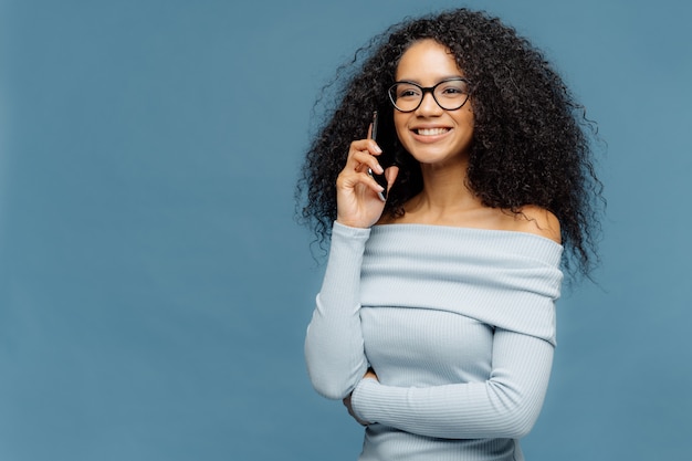 Sonriente mujer afroamericana encantada con el pelo rizado, habla por teléfono inteligente