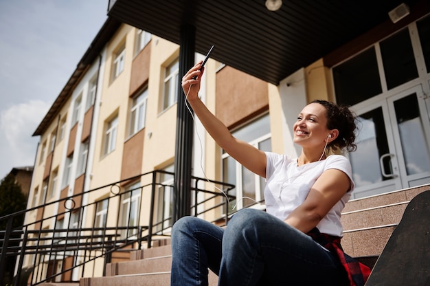 Sonriente mujer afroamericana feliz con auriculares en ropa casual haciendo selfie mientras está sentado en los pasos en el fondo del edificio amarillo