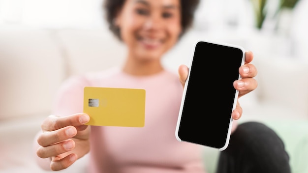 Sonriente mujer afroamericana milenaria muestra tarjeta de crédito y teléfono inteligente con pantalla en blanco en la sala de estar