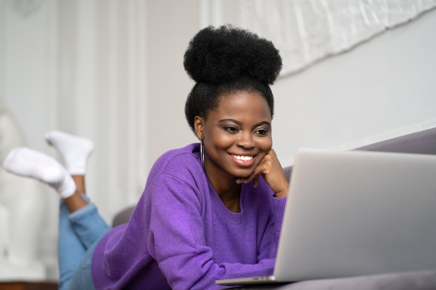 Sonriente mujer afroamericana con peinado afro usar suéter púrpura acostado en el sofá, descansando, mirando la webcam