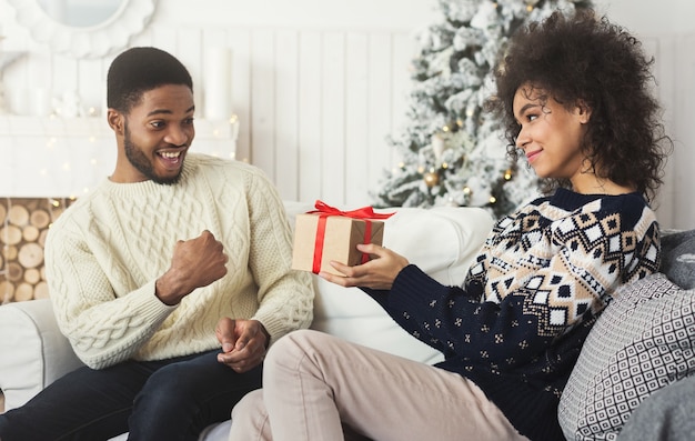 Sonriente mujer afroamericana que presenta un regalo al novio sorprendido mientras celebra la Navidad en casa