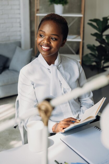 Sonriente mujer afroamericana sosteniendo un cuaderno en las manos pensando en tomar notas trabajando desde su nueva...