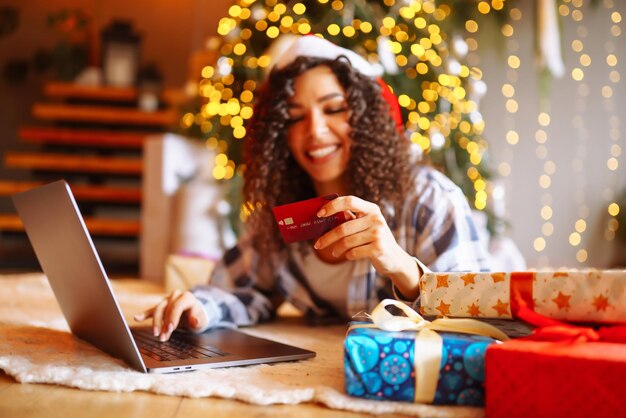 Sonriente mujer afroamericana usando una laptop sentada cerca del árbol de Navidad con tarjetas de compras en línea