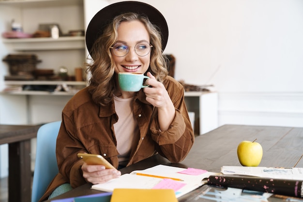 Sonriente mujer agradable en anteojos y sombrero con teléfono celular mientras bebe café en casa