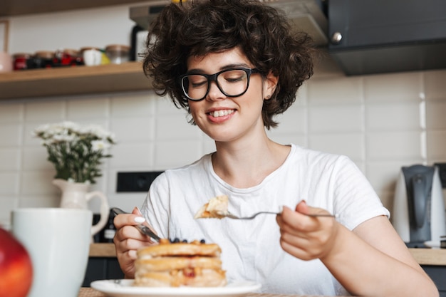 Sonriente mujer alegre desayunando sabroso mientras está sentado en la cocina en casa, comiendo panqueques