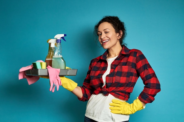 Sonriente mujer americana posando en azul con bandeja con productos de limpieza en las manos y una mano en la cintura.