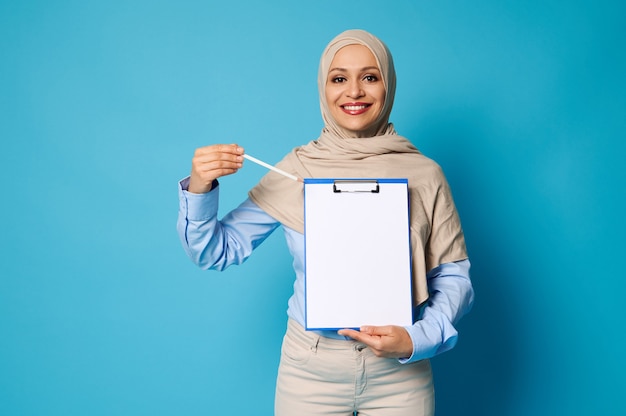 Sonriente mujer árabe con la cabeza cubierta en hijab apuntando con un bolígrafo sobre una superficie en blanco sobre una hoja de papel blanco en el portapapeles