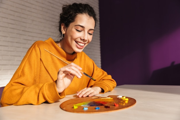 Sonriente mujer artística eligiendo el color de la pintura en una paleta en el interior