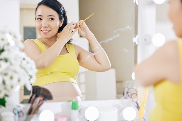 Sonriente mujer asiática bastante embarazada de pie en el espejo en el baño y cepillarse el pelo