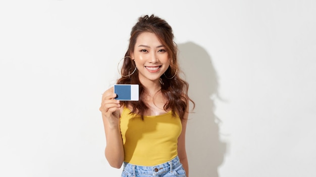 Sonriente mujer asiática bonita sosteniendo una tarjeta de crédito mientras se alegra y mira a la cámara sobre un fondo blanco